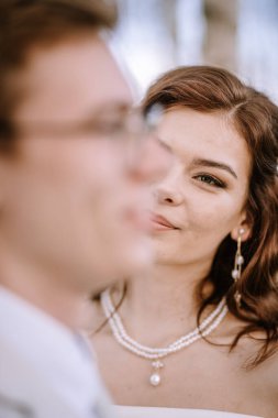 Valmiera, Letonya - 9 Eylül 2023 - Bir gelinin damadına yumuşak bir gülümsemeyle bakması, kolyesini ve küpelerini orman arka planında göstermesi.