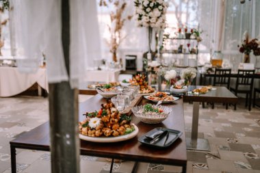 Valmiera, Letonya - 9 Eylül 2023 - Çiçek düzenlemeleri ve yumuşak doğal ışıkla süslenmiş zarif bir odada çeşitli tabak ve cam örtülerle dolu bir açık büfe masası.