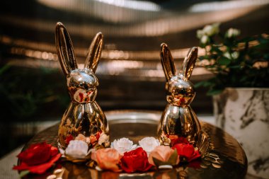 Valmiera, Letonya - 9 Eylül 2023 - Kırmızı ve beyaz güllerle süslenmiş iki altın tavşan heykeli, arka planda bulanık ışıklar bulunan yansıtıcı bir yüzeye kurulmuş.