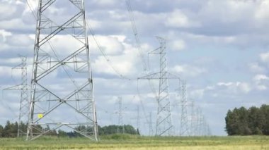 Yüksek voltajlı elektrik hattı kuleleri kırsal bir arazi boyunca uzanır. Mavi gökyüzünün altındaki tarlalar ve ağaçlarla sıcak havada dağınık bulutlar..
