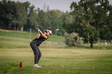 Sigulda, Letonya - 10 Eylül 2023 - Siyah elbiseli kadın golfçü yeşil bir golf sahasında sallanıyor, arkasında ağaçlar ve kırmızı bir işaret var.