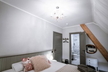 Blome, Letonya - 11 Eylül 2023 - Düzgün yapılmış yatağı, çiçekli ve kürklü yastıkları, avizesi, katlanmış havluları, komodini ve modern bir banyo kapısını olan rahat bir yatak odası.
