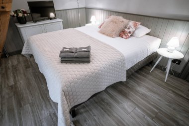 Blome, Letonya - 11 Eylül 2023 - Düzgün yapılmış yatağı, çiçekli ve kürklü yastıkları, lambalı komodinleri, yatağında katlanmış havluları ve dolabında TV olan rahat bir yatak odası.