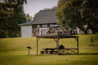 Blome, Letonya - 11 Eylül 2023 - Piknik masalı ahşap çardak ve arka planda kır evi ve bahçesi olan çimenli bir tepede ızgara. Boşluğu kopyala.