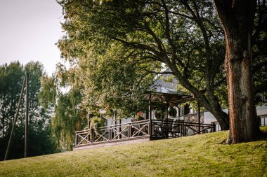 Blome, Letonya - 11 Eylül 2023 - Güneşli bir günde yemyeşil ağaçlar ve yeşilliklerle çevrili ahşap güverteli büyüleyici kır evi. Boşluğu kopyala.