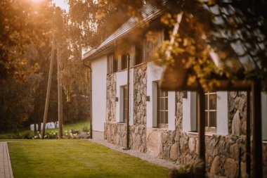 Blome, Letonya - 11 Eylül 2023 - Taş ve sıva evin dış tarafı geniş pencereler ve yemyeşil çimlerle kaplı, sıcak güneş ışığı altında, ağaçlar ve yeşilliklerle çevrili.