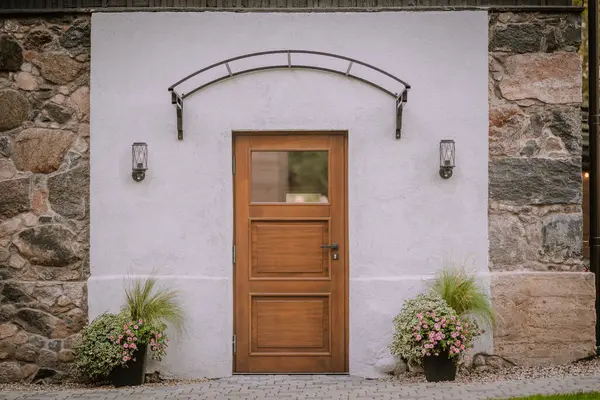 stock image Blome, Latvia - September 11, 2023 - A wooden door with a window, flanked by two potted plants, set in a white and stone wall, featuring two wall-mounted lanterns.