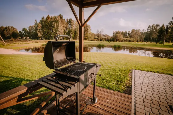 stock image Blome, Latvia - September 11, 2023 - An outdoor grill with an open lid on a wooden deck, overlooking a serene pond and grassy landscape in a park setting.