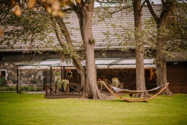 Blome, Letonya - 11 Eylül 2023 - Yeşil çimenlikteki taştan bir evin yanında asılmış bitkilerle ve ağaçlar arasında bir hamağıyla rahat bir açık hava çardağı.