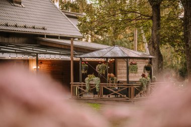 Blome, Letonya - 11 Eylül 2023 - Çevresi ağaçlar ve yeşilliklerle çevrili kırsal bir ahşap evin bitişiğinde asılı çiçek saksıları ve ışıkları olan rahat açık hava çardağı.