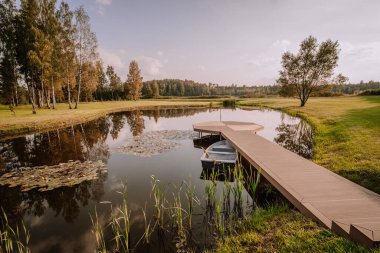 Blome, Letonya - 11 Eylül 2023 - Nilüfer yapraklı sakin bir gölette kayıkla tahta iskele, ağaçlarla çevrili ve parçalı bulutlu bir gökyüzünün altında çimenli bir tarla.