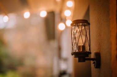 Blome, Letonya - 11 Eylül 2023 - Arka planda parlayan sicim lambaları olan kırsal bir duvar fenerinin yakın çekimi, sıcak ve davetkar bir atmosfer yaratıyor. Boşluğu kopyala.