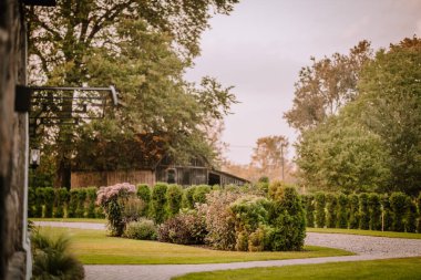 Blome, Letonya - 11 Eylül 2023 - yemyeşil çalıları ve ağaçları olan güzel bir bahçe, bir çakıl yolu ve arka planda kırsal bir ahşap bina. Boşluğu kopyala.