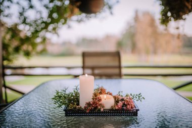 Blome, Letonya - 11 Eylül 2023 - Çiçekler ve yeşillikle süslenmiş cam bir masanın üzerinde iki mum, bulanık bir arka fona karşı kurulmuş. Boşluğu kopyala.