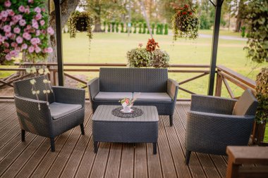 Blome, Letonya - 11 Eylül 2023 - Hasır mobilyaları, asılı bitkileri ve güneşli bir günde yeşilliklerle çevrili küçük bir masası olan bir balkonda açık hava oturma alanı. Boşluğu kopyala.