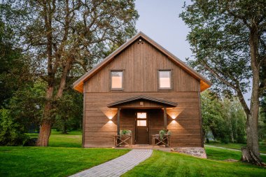 Blome, Letonya - 11 Eylül 2023 - Sıcak ışıkları ve taş temeli olan ahşap bir kulübenin ön görüntüsü, yemyeşil çimenler ve kapıya giden tuğla yolu olan uzun ağaçlar arasında geçmektedir..