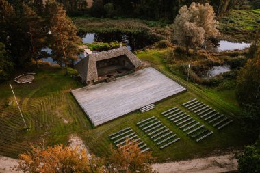 Blome, Latvia - September 11, 2023 - Aerial view of an outdoor amphitheater with wooden benches and a stage, surrounded by greenery and a small pond. clipart