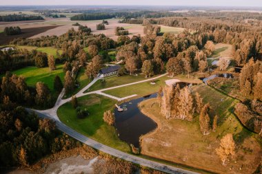 Blome, Letonya - 11 Eylül 2023 - Güneşli bir günde, bir ev, gölet, çardak ve çevreleyen ağaçlar, tarlalar ve yollar ile birlikte kırsal bir arazinin havadan görünüşü,.