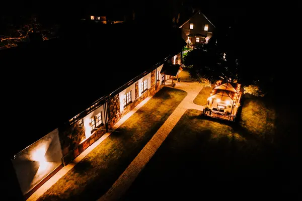 stock image Blome, Latvia - September 11, 2023 - Aerial night view of an illuminated stone house and a well-lit gazebo with surrounding garden paths and trees, creating a cozy and inviting ambiance.