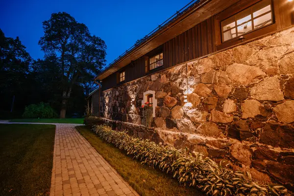 stock image Blome, Latvia - September 11, 2023 - Stone wall of a building illuminated at night with path and garden plants in the foreground, creating a warm and inviting atmosphere.