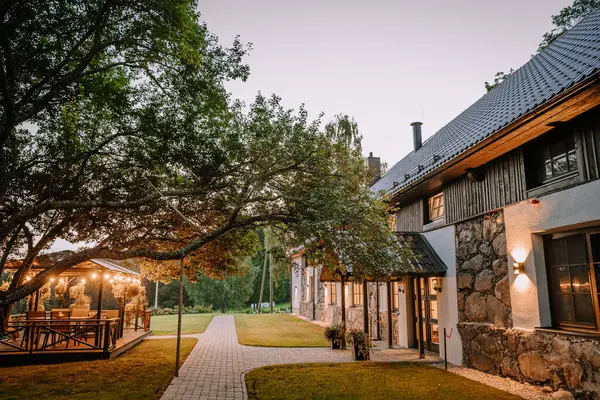 stock image Blome, Latvia - September 11, 2023 - Cozy stone and wood house with illuminated outdoor seating area under a large tree, lit by warm lights, creating a serene evening ambiance.