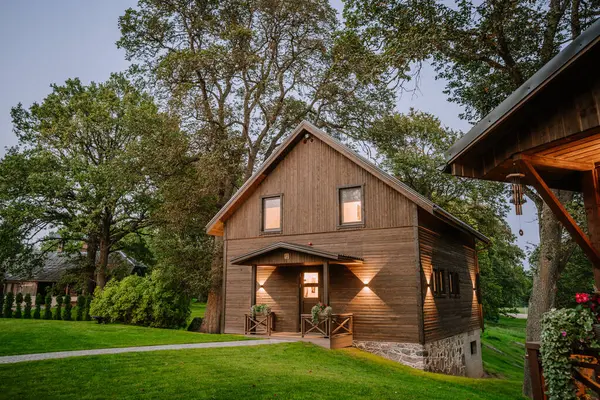 stock image Blome, Latvia - September 11, 2023 - A wooden cabin with illuminated exterior, surrounded by lush greenery, trees, and paved walkways in a peaceful setting during dusk.