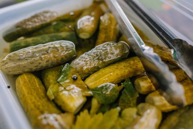 limbazi, Letonya, 29 Haziran 2024 - Maşa dolu bir konteynırda turşu yakın çekim, tuzlu suya batırılmış ve baharatlarla süslenmiş, servis için hazır.