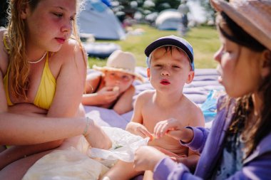 limbazi, Letonya, 29 Haziran 2024 - Çocuklar kamp alanında bir şişme yatakta oturur, abur cubur yer ve arka planda çadırlar ve yeşilliklerle güneşli bir günün tadını çıkarırlar.