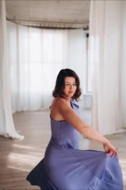 Lavanta elbiseli bir kadın, beyaz perdeleri ve doğal ışığı olan ferah, parlak bir odada güvenle poz veriyor. Boşluğu kopyala.
