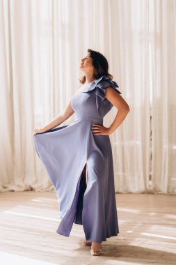 Lavanta elbiseli bir kadın beyaz perdeleri ve doğal ışığı olan parlak bir odada eteği kaldırıp tutuyor. Boşluğu kopyala.