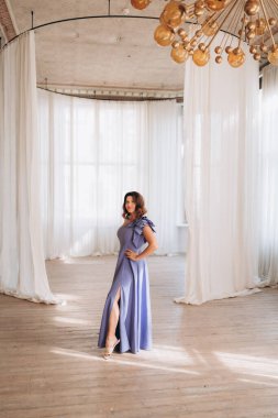  Lavanta elbiseli zarif bir kadın. Geniş, parlak bir odada poz veriyor. Beyaz perdeleri ve modern bir avizesi var..
