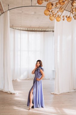  Lavanta elbiseli zarif bir kadın. Geniş, parlak bir odada poz veriyor. Beyaz perdeleri ve modern bir avizesi var..