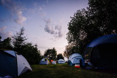 Limbazi, Letonya - 28 Haziran 2024 - Mor ve mavi gökyüzünün altındaki ağaçlarla çevrili çimenli bir alanda çadırlarla kurulmuş akşam kampı sahnesi. Boşluğu kopyala.