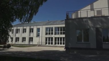 Gulbene, Letonya - 28 Mayıs 2024 - Geniş pencereli, açık gri cepheli ve önünde açık mavi gökyüzüne karşı duran bankları olan modern bir binanın videosu.