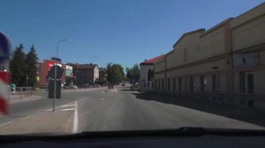 Balvi, Letonya - 28 Mayıs 2024 - Açık mavi gökyüzü altında çeşitli mağazaları ve binaları olan bir şehir sokağının videosu, bir arabanın gösterge panelinden alındı.
