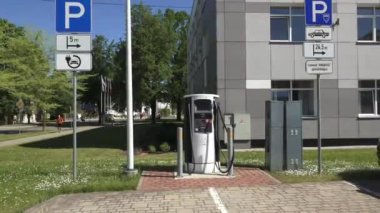 Balvi, Letonya - 28 Mayıs 2024 - Güneşli bir günde modern bir binanın dışındaki park işaretli ve yeşillikli elektrikli araç şarj istasyonunun videosu. Boşluğu kopyala.