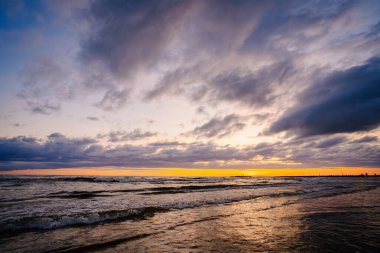 Okyanusun üzerinde dramatik bir günbatımı dalgalar ve karanlık, dağınık bulutlarla dolu bir gökyüzü karamsar ve karamsar bir sahne yaratıyor..