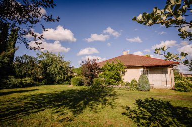 Valmiera, Letonya - 5 Temmuz 2024 - Güneşli bir günde açık mavi gökyüzünün altında çeşitli ağaçlar, çalılar ve yeşil çimlerle çevrili kırmızı çatılı bir ev.