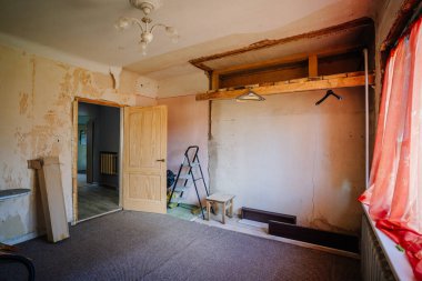 Valmiera, Letonya - 5 Temmuz - 5 Temmuz 2024 - Tavan lambası armatürünün altında açıkta kalmış kirişler, soyulmuş duvar kağıtları, ahşap bir kapı, bir merdiven ve kırmızı perdelerle dolu tamamlanmamış bir oda.