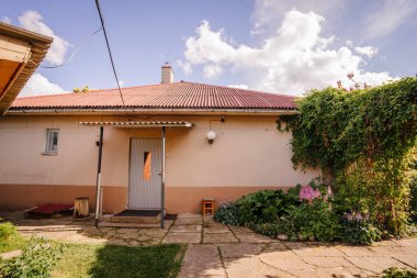 Valmiera, Letonya - 5 Temmuz 2024 - Kırmızı çatılı ve küçük verandalı, yemyeşil ve çiçeklerle çevrili, bulutlu açık mavi bir gökyüzünün altında.