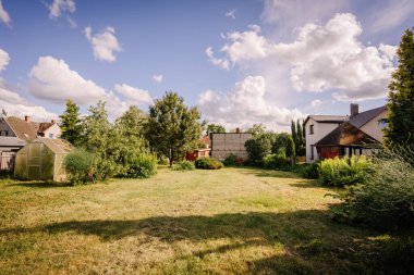Valmiera, Letonya - Temmuz - 5, 2024 Tüylü bulutlu mavi gökyüzü altında serası, ağaçları ve evleri olan geniş bir arka bahçe..