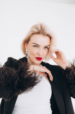 Kırmızı rujlu, beyan küpeli, tüylü siyah ceketli, düşünceli bir gülümsemeyle poz veren sarışın bir kadın..