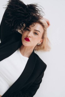 Tüylü manşetli, beyaz bluzlu, siyah ceketli bir kadın yerde uzanmış, kendinden emin bir ifadeyle kameraya bakıyor..