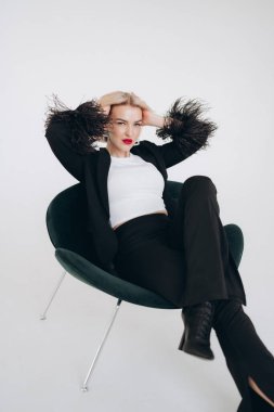 Siyah ceketli, tüylü ve siyah pantolonlu şık bir kadın yeşil bir sandalyede uzanır, kendine güveni ve entelektüelliği vardır..