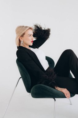 Siyah ceketli şık, sarışın bir kadın. Tüylü kolları yeşil bir sandalyede uzanıyor. Minimum bir arka plana karşı özgüven ve zarafet yayıyor..