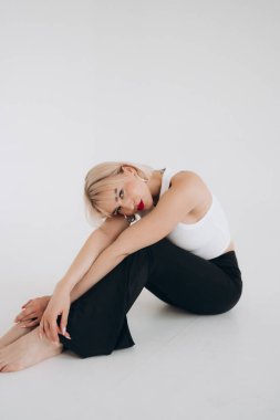 Beyaz kesim bluzlu ve siyah pantolonlu sarışın bir kadın başını dizlerinin üzerine yaslamış, düz beyaz bir arka plana yaslanmış şekilde yerde oturuyor..