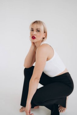 Beyaz bluzlu, siyah pantolonlu sarışın bir kadın yere diz çöker, yan tarafa bakar, düz beyaz bir arka plana bakar..