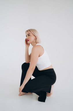 Beyaz bluzlu, siyah pantolonlu sarışın bir kadın yere diz çöker, yan tarafa bakar, düz beyaz bir arka plana bakar..