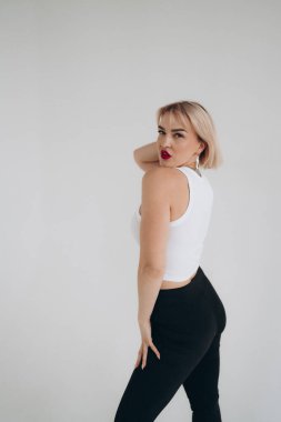 Beyaz bluzlu ve siyah pantolonlu sarışın bir kadın kolları yukarı doğru uzanmış, düz beyaz bir arkaplan üzerine kurulmuş zarif poz veriyor..