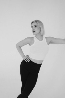 Beyaz bluzlu ve siyah pantolonlu sarışın bir kadın düz beyaz bir arka plana yaslanmış bir duvara yaslanmış bir şekilde poz veriyor..